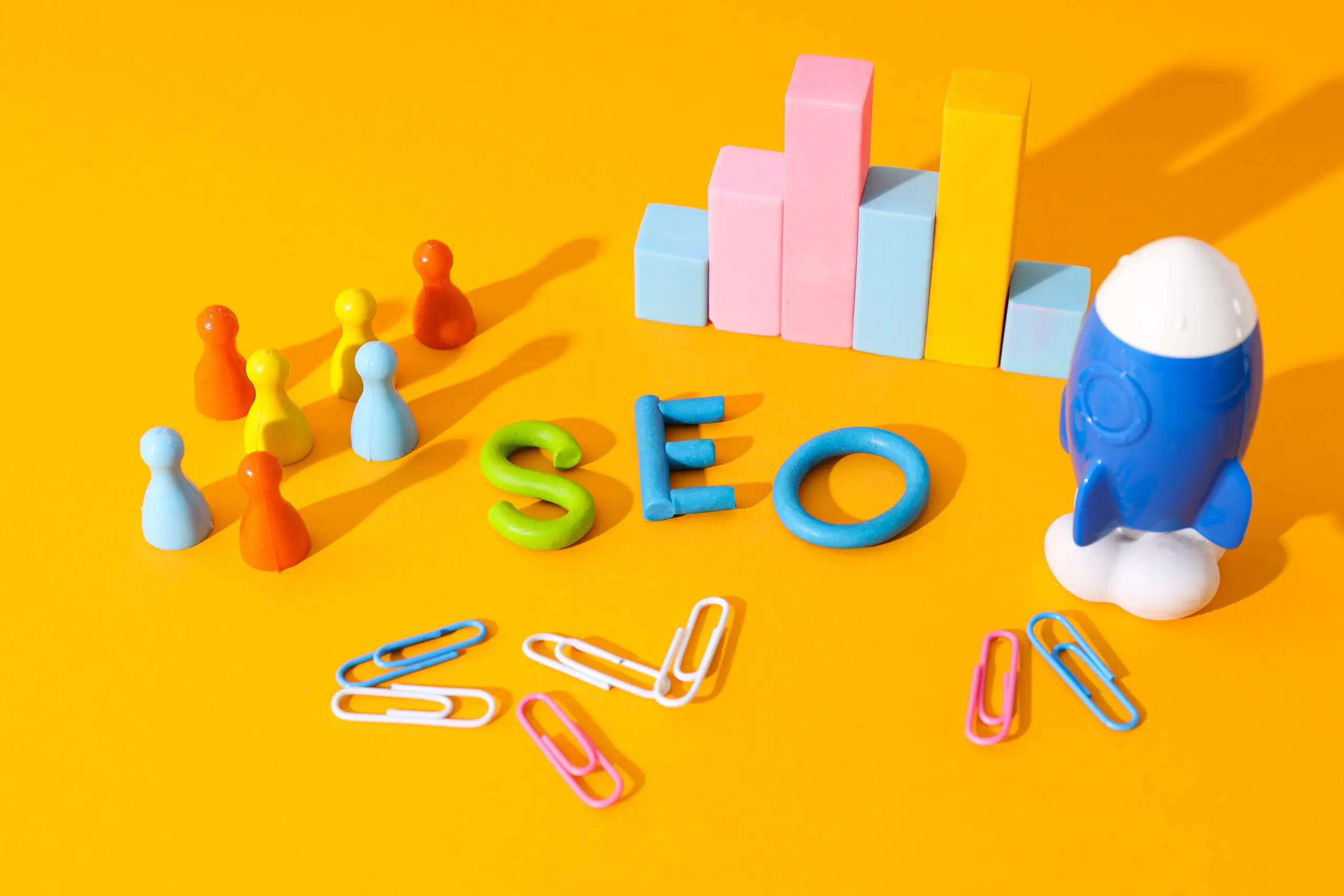 Colorful SEO letters, a group of multicolored game pieces, a blue toy rocket, and a graph made of blocks symbolize Local SEO strategies, all neatly arranged with paperclips on a bright yellow background. Colorful SEO letters, a group of multicolored game pieces, a blue toy rocket, and a graph made of blocks symbolize Local SEO strategies, all neatly arranged with paperclips on a bright yellow background.